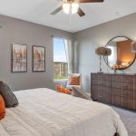 A modern bedroom with a neatly made bed featuring orange and neutral pillows, two large windows, a dresser with a round mirror, decorative lamps, wall art, and a potted plant on beige carpet.