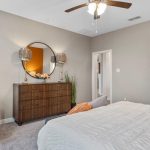 A modern bedroom with a neatly made bed, gray and orange accent pillows, a wooden dresser with a round mirror, two lamps, a white chair, beige walls, and a ceiling fan with lights.