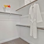 A spacious walk-in closet with white walls, empty wooden hanging rods, a white robe hanging on one rod, a white shelf with neatly folded towels, and a white shopping bag with red and yellow tissue paper.