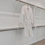 A white bathrobe hangs on a wooden rod in an empty walk-in closet with white walls, multiple shelves, and carpeted flooring. A plastic bin with items sits on the upper right shelf.