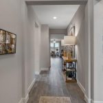 A modern hallway with gray walls and wood flooring leads to a bright living area; a bedroom is visible to the right, and a console table with a lamp and decor is along the hallway. Abstract art hangs on the left wall.