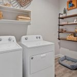A bright laundry room with a white washer and dryer, shelves holding baskets and towels, and a corner shelving unit with pet supplies above a dog bed on the floor. A framed dog portrait hangs on the wall.