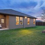 A single-story house with beige siding and large windows sits on a well-kept green lawn, with a small patio area, a young tree, and a wooden fence under a blue and pink sunset sky.