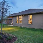 A single-story tan house with large windows, a well-kept lawn, a small tree, and landscaped garden beds, photographed at dusk with interior lights glowing.