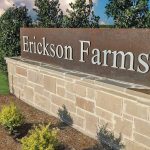 A stone and metal entrance sign reads "Erickson Farms," surrounded by green shrubs, grass, and trees, with a stone wall extending into the background.