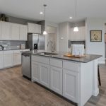 Modern kitchen with light gray cabinets, stainless steel appliances, a central island with sink, and wood-style flooring. Walls are painted light gray, and the space is well-lit with recessed and pendant lighting.
