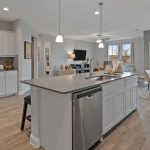 Modern, open-concept kitchen with white cabinets, a large island with sink and dishwasher, pendant lighting, adjacent dining area, and living room with TV; large windows provide natural light.
