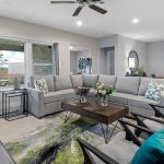 Modern living room with gray sectional sofa, two dark armchairs with teal pillows, a wooden coffee table with flowers, and sliding glass doors leading to a patio. A dining area with green chairs is visible in the background.