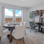 A bright home office with a round table and four upholstered chairs, a desk with a rolling chair, bookshelves, modern decor, framed leaf art, and a window with patterned curtains letting in natural light.
