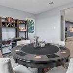 Modern dining area with a round black table and four upholstered chairs. A desk with books and artworks featuring green palm leaves are against a gray wall. Bright, open living space is visible in the background.