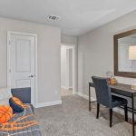 A cozy room with a gray carpet, a small desk and chair, a large mirror, a lamp, and a daybed decorated with pillows and a basketball. The walls are light beige and two doors are visible.