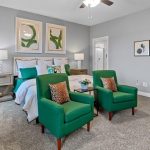 A modern bedroom with light gray walls, a bed with blue and white bedding, two green armchairs with patterned pillows, bedside tables with lamps, framed art on the walls, and beige carpet flooring.