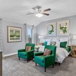 A modern bedroom with gray walls, a bed with white and green bedding, two green chairs at the foot, wooden nightstands, lamps, large windows, and abstract art on the walls. A ceiling fan hangs above.