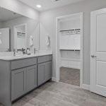 Modern bathroom with gray double-sink vanity, large mirror, and white walls; open door leads to a walk-in closet with shelves and hanging rods. Towels hang beside the mirror, and a landscape painting is on the wall.