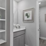 A modern bathroom with gray cabinets, a white sink, open shelves with towels and toiletries, framed abstract art on the walls, and a toilet near the back. The space is clean and well-organized.