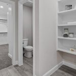 A hallway with built-in white shelves holding towels, jars, and small boxes. To the left, a doorway leads to a bathroom with a toilet and a walk-in closet with white cabinetry, hangers, and gray carpet.