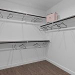 Empty walk-in closet with white walls, brown carpet, several black hangers on metal rods, and a pink storage box on the top shelf.