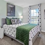A modern bedroom with a green and white polka dot bedspread, two bedside tables with lamps, ballerina artwork on the walls, a window with patterned curtains, and neutral-toned carpet.