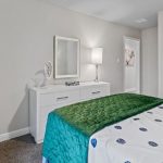 A modern bedroom with a white dresser and mirror, a green blanket on the bed, light-colored walls, and a closed white door. A small decorative item and a lamp sit on the dresser.