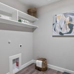 A modern laundry room with light gray walls, open shelves holding baskets and towels, abstract wall art, and a laundry basket on the floor next to a pillow labeled "Reserved.