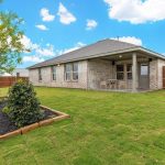 A spacious backyard with green grass, a young tree, small shrubs, a covered patio area with chairs, a light brick house, and a wooden privacy fence under a blue sky with scattered clouds.
