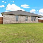 A brick house with a dark roof sits in a spacious backyard with a green lawn, surrounded by a wooden privacy fence under a blue sky with scattered clouds.