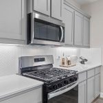 Modern kitchen with light gray cabinets, stainless steel stove and microwave, white countertops, and a white herringbone tile backsplash. A cookbook and utensils sit on the counter next to the stove.