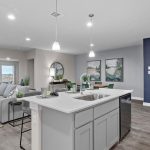 Modern open-concept living space with a kitchen island in the foreground, gray sectional sofa, dining area, wall art, and large windows letting in natural light. Decor is sleek with neutral tones and pops of green.