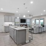 Modern open-concept kitchen and living room with light gray cabinets, an island with stools, stainless steel appliances, a sectional sofa, large windows, and wood-look flooring. Dining area visible in the background.