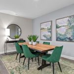 A modern dining room with a wooden table, six green chairs, a green-patterned rug, two landscape paintings on a gray wall, a console table with a round mirror, and potted plants as decor.