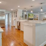 Modern kitchen with white cabinets, marble countertops, stainless steel appliances, and wood flooring. Pendant lights hang over a large island. An open layout leads to a living area and a staircase in the background.