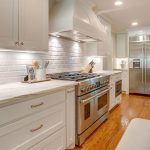 Modern kitchen with white cabinets, stainless steel double oven and refrigerator, marble countertops, white subway tile backsplash, hardwood floors, and under-cabinet lighting. Kitchen utensils and decor are on the counters.