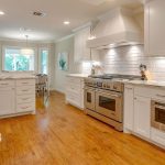 Bright, modern kitchen with white cabinets, marble countertops, stainless steel appliances, a gas range, and wood floors. A dining area with large windows and a round table is visible in the background.