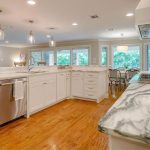 Bright, modern kitchen with white cabinets, marble countertops, stainless steel appliances, wooden floors, and pendant lighting. Large windows and a dining area with chairs are visible in the background.