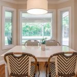 A bright dining area with a white table, four patterned chairs, a potted plant centerpiece, large windows showing a green backyard, and a round overhead light fixture. Abstract art hangs on the right wall.