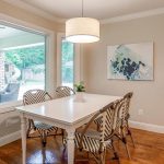 A dining area with a white table and four patterned chairs, hardwood floors, a modern pendant light, abstract wall art, and large windows overlooking a patio and green yard.