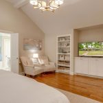 A spacious, well-lit bedroom with wooden floors, a white bed, two beige armchairs, built-in shelves, and a TV. The room connects to a bathroom and an open hallway leading to another room.