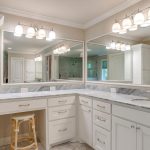 Bright, spacious bathroom with white cabinetry, marble countertops, two large mirrors, modern light fixtures, a single sink, and a wooden stool tucked under the counter. Reflections show more of the room.