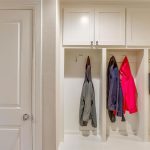 A mudroom with white built-in cubbies, each with hooks. Two cubbies hold jackets, and one holds a bright pink coat and a patterned tote bag. A closed white door is on the left.