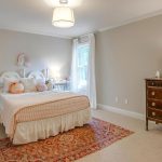 A cozy bedroom with a white bed, peach plaid bedding, plush toys, a nightstand, a window with white curtains, a wooden dresser with a lamp and framed art, and a patterned rug on a beige carpet.