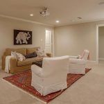 A cozy living room with beige walls, a sofa, two white armchairs on a patterned rug, a bookshelf, and elephant wall art. There is a ceiling projector and closed double doors in the room.