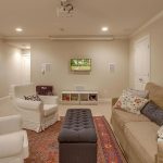 A cozy living room with beige walls, a tan sofa, two white armchairs, a patterned rug, a dark ottoman, a small TV on the wall, and an elephant artwork above the couch. The room has soft, warm lighting.