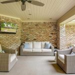A covered patio with brick walls features a wicker sofa and two matching armchairs with white cushions, a ceiling fan, a mounted TV, and a view of a grassy backyard with trees.