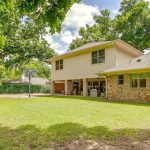 A two-story house with brick and siding, seen from the backyard. The yard features green grass, a basketball hoop, a swing set, and is surrounded by trees and a wooden fence.