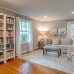 A cozy living room with bookshelves full of books, a light gray sectional sofa with pillows, a cushioned bench, side tables, lamps, and windows with white curtains. Hardwood floors and soft lighting create a warm atmosphere.