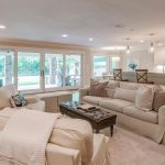 Spacious, open-concept living room and kitchen with neutral-toned sofas, a tufted ottoman, bar stools at a white kitchen counter, large windows, and sliding glass doors overlooking a green backyard.