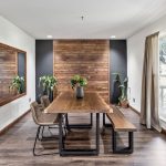 A modern dining room with a wooden table, two chairs, and a matching bench. The room features wood paneling on the far wall, a large mirror, potted plants, and a window with beige curtains letting in natural light.