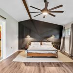 A modern bedroom with a geometric black accent wall, a wooden bed with white bedding, two wall lamps, wood flooring, a beige rug, large ceiling fan, sliding barn door, and windows with gray curtains.