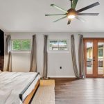 Modern bedroom with a large bed, dark accent wall, and a ceiling fan. Two windows and a glass door with gray curtains let in natural light. A geometric mirror and potted plant sit by the door on dark wood floors.