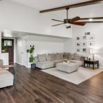 A modern living room with wood floors, white walls, exposed ceiling beams, a large sectional sofa, coffee table, floor lamp, framed photos on the wall, and a ceiling fan. A green plant sits near a partial brick wall.